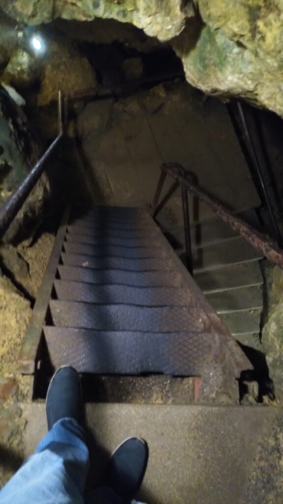 Symbolbild für das Innenweltreisen: Füße am oberen Absatz einer Treppe aus Metall und Beton die in einer Höhle nach unten führt.