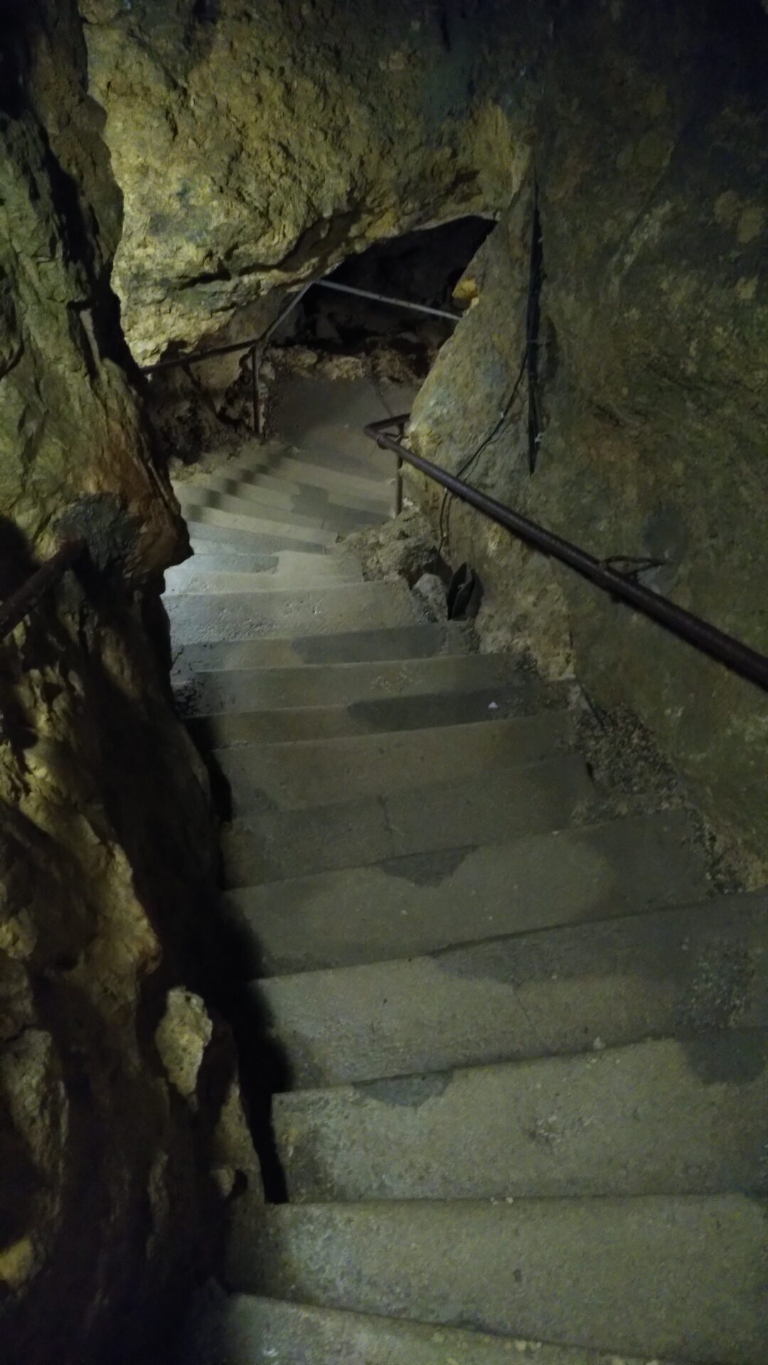 Symbolisch für den Weg ins eigene Unterbewusstsein: Eine Treppe die nach unten führt. Betonstufen, ein Handlauf aus Metall, die Wände teils roher teils behauener Fels.